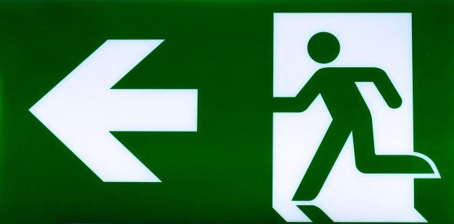 An Exit sign with a green field & man running out a door & a white arrow pointing to the left.